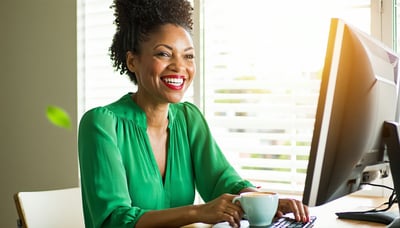 A cheerful woman with a radiant smile sits comfortably at her desk adorned in a vibrant green blouse that accentuates her lively personality As she types away on her computer she takes a moment to savor a steaming cup of coffee the rich aroma wafting